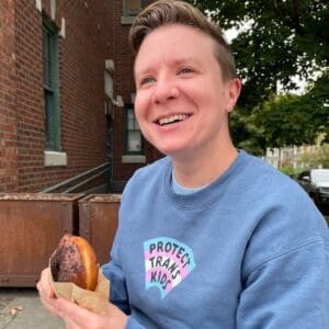 Non-gender conforming person smiling and eating a bagel while wearing a blue sweatshirt that reads "protect trans kids"