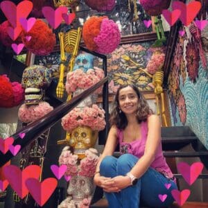 Smiling woman wearing jeans and a pink shirt seating in front of a day of the dead alter with hearts imprinted on the photo