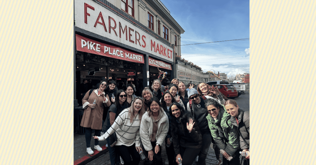 17 women smiling at Pike Place Market under a blue sky