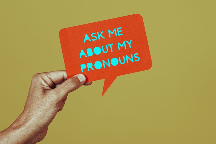 A hand holds a sign that says, “Ask me about my pronouns.”